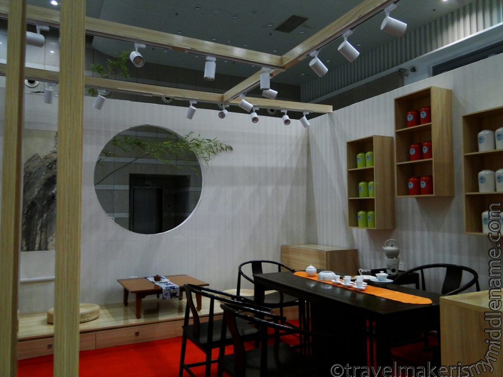A style stall at the Spring Xiamen Tea Fair with traditional Chinese furniture: a long wooden table with wooden chairs with arm and back rests, and a round window in the wall.