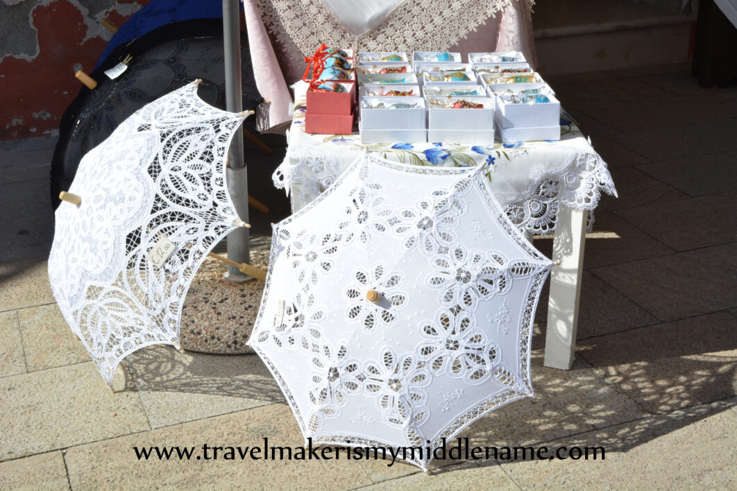 two white lace parasols on the stone ground for sale in Burano, Venice, Italy. A small table with rows of boxes of glassware in the background. Photo copyright and credit: Author