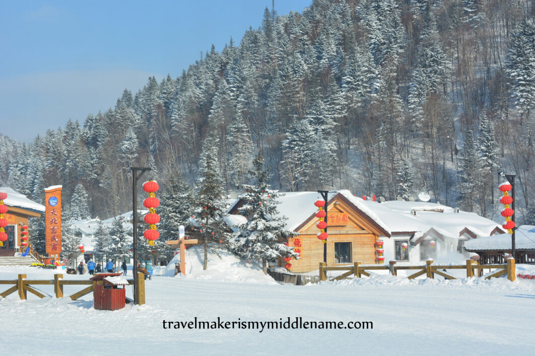 Daytime: A timber building covered in snow on a white, snow covered ground, with several red lanterns hanging in a column from street lamps and a forested mountain dusted with snow in the background.