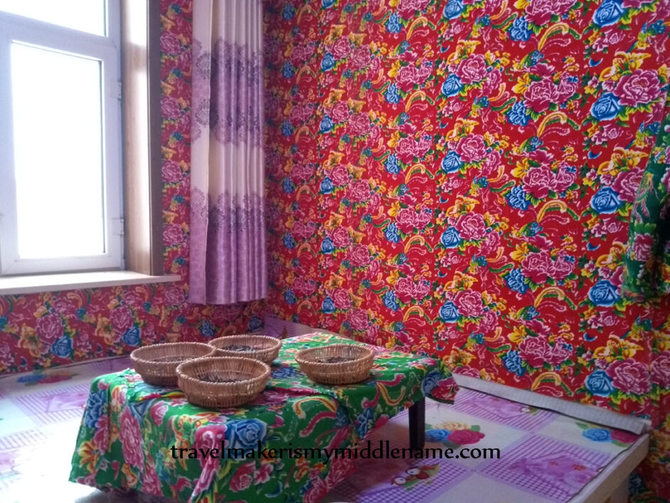 Daytime, interior: A display home showing a room inside a northern Chinese style house with a kang bed and decorated in the northern Chinese trademark colourful floral design. A small table facing the window sits on the kang bed and has some snacks on it.