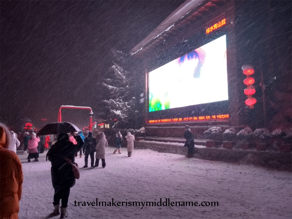 Night time, exterior: A giant LED screen lights up the snow covered grounds on the main street where people are walking in Snow Town, China. Snow is falling and one person has an umbrella.