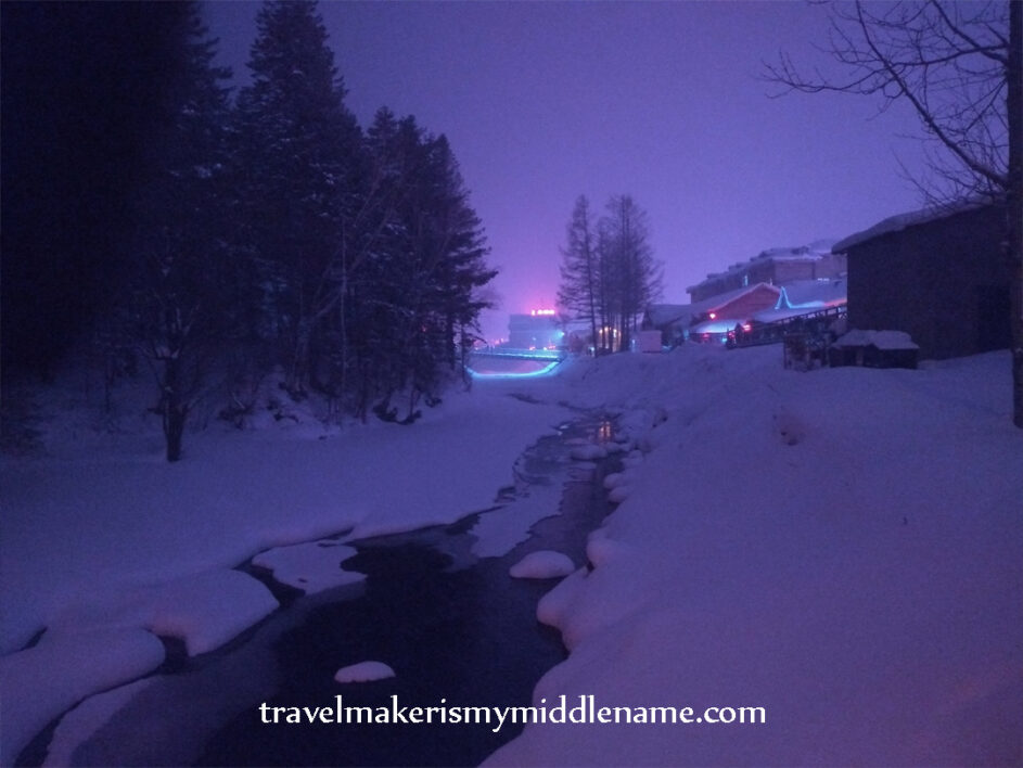 Night time, exterior: a small creek flows through the town despite the temperature being minus 30 degrees Celsius with snow on either side and trees to one side. Lights of the town in the distance can be seen. 