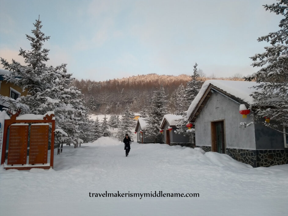 The sun shines onto the tip of the mountain behind a residential area at sunrise. A person walks through the snow outside their hotel. 