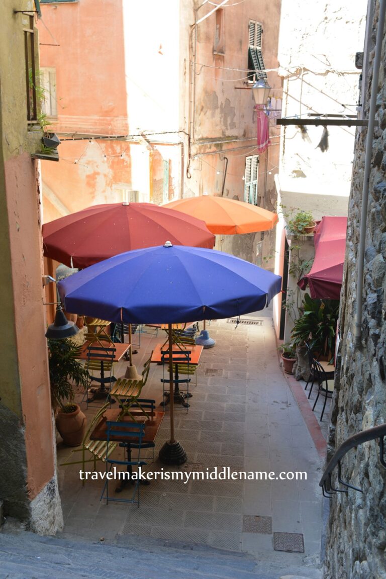 Three umbrellas, orange, deep pink and navy blue cover me outdoor table and chairs in Vernazza in a an alleyway space between buildings.