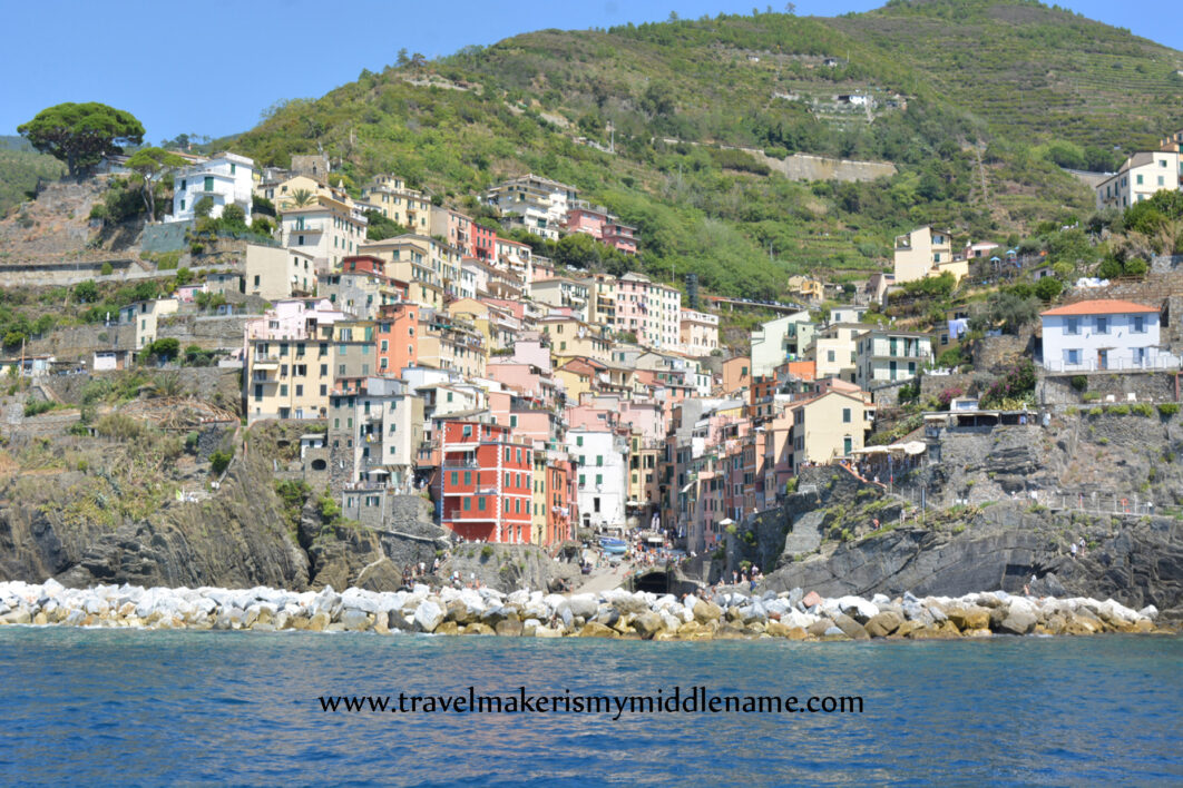 A wide shot of Riomaggiore in Cinque Terre in Italy shot from the ocean showing the buildings on the cliffs. In the background are the green terraces for planting and a cloudless blue summer sky. Part of the dark blue ocean is seen in the foreground.