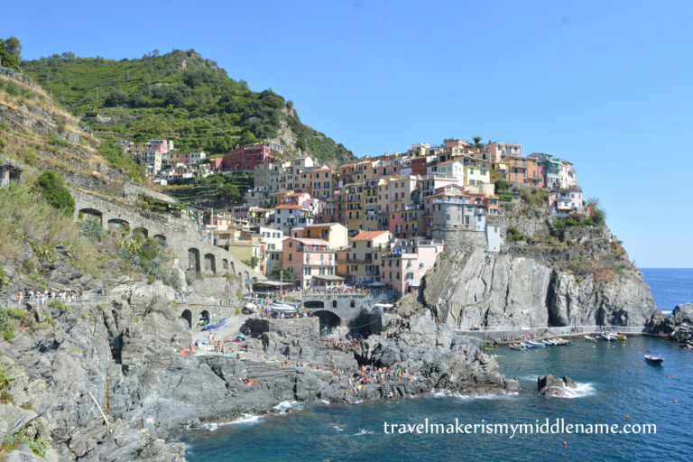 A wide shot of Riomaggiore in Cinque Terre in Italy showing the buildings on the cliffs. In the background are the green terraces for planting and a cloudless blue summer sky. Part of the dark blue ocean is seen in the foreground.