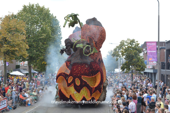 A front view of a giant smoking jack-o-lantern parade float in the middle of the street with trees and crowds on either side against a white sky.
