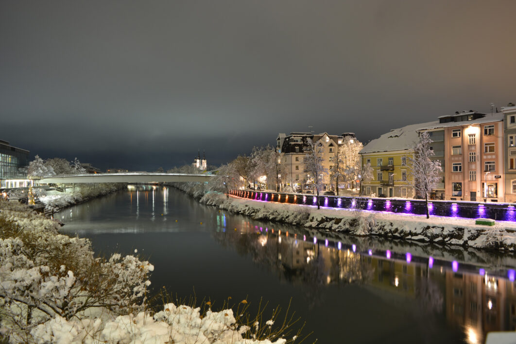 The Drava River in Austria at night as it curves around the street. A bridge is in the far distance, on the left are snow covered branches, on the right are buildings along the street and purple lights that line the street.