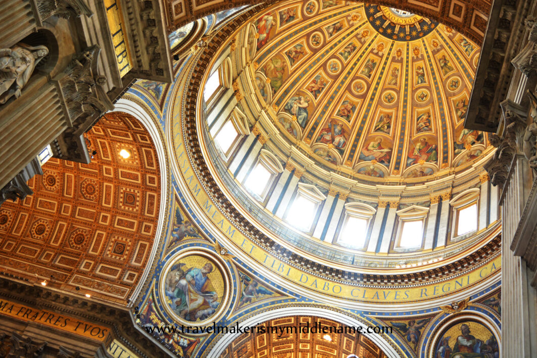 An upwards view of the giant, painted dome inside St Peter's Basilica in the Vatican City, Rom, Italy. Light pours in the windows of the dome and giant archways can be seen on the left and bottom middle of the image, all covered in orangey gold paint.