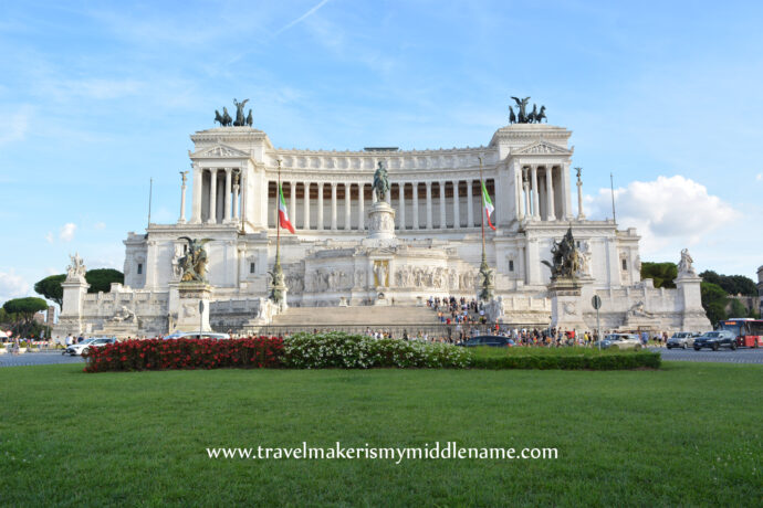 A front view of the Vittoriano in Rome, Italy. during the day with a blue cloudless sky in the back and green grass in the foreground. A shrub with plants in colours of the Italian flag, from left to right: red, white and green is on the grassy field.