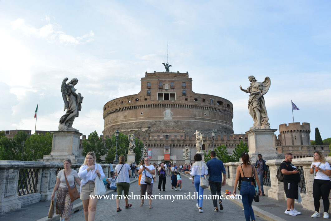 A front view of the Castel Sant'Angelo seen from the street end of the bridge. An angle sculpture is on either side of the bridge. People walk forwards and back on the bridge between the street and castle.