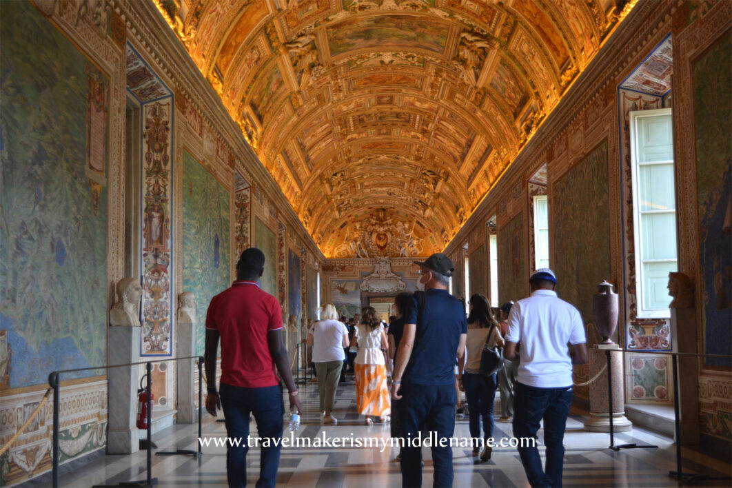 A view of hallway of the Vatican Museum with the long, curved, narrow ceiling seen from one end. People walk down the hallway away from the view and windows on the right hand side light up the call. A wall with artwork on the left side.