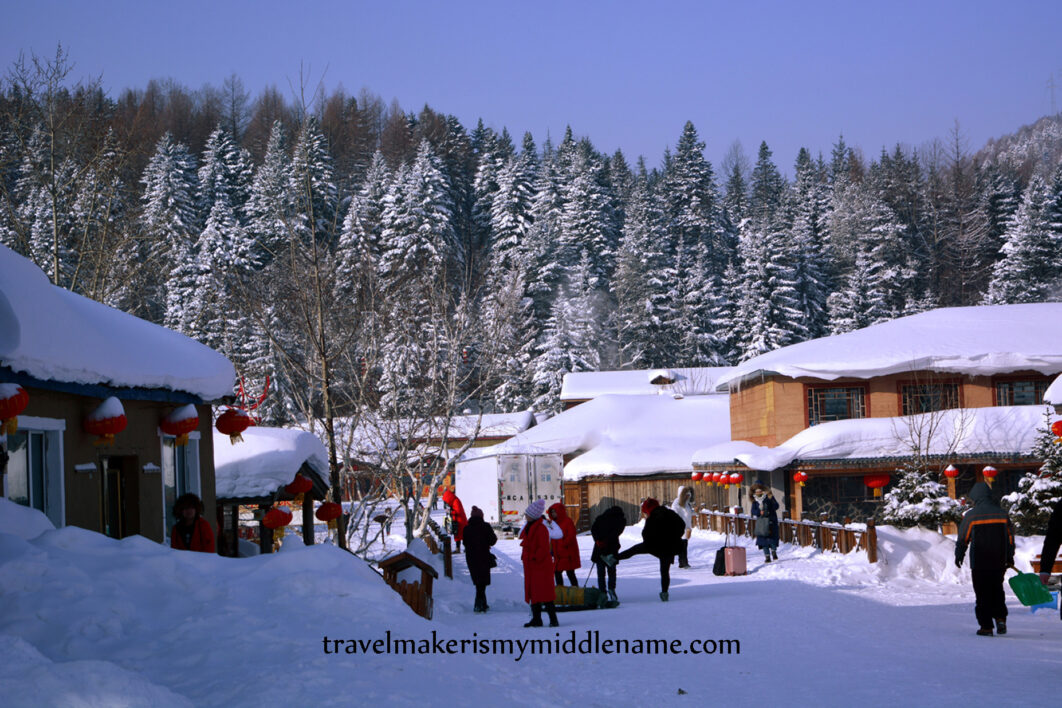 Daytime: Snow covered buildings with a forest covered mountain behind it. A few people walk through the snow covered grounds as a delivery truck slowly moves ahead.