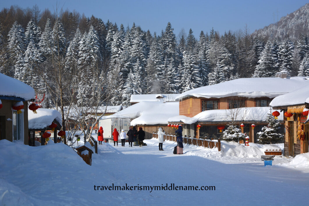 Daytime: Snow covered buildings with a forest covered mountain behind it. A few people walk through the snow covered grounds.