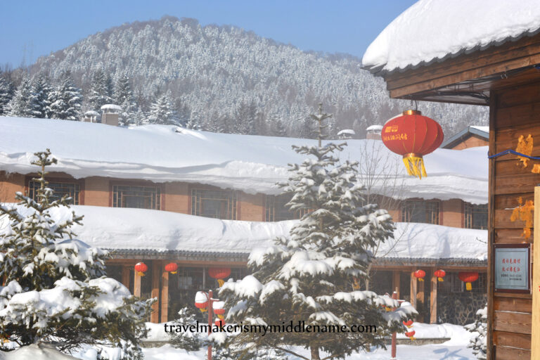 Daytime: Snow covered buildings with red lanterns dangling from the roof with forested mountains in the background.