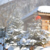 Daytime: A red lantern dangles from the roof of a timber house with snow covered trees in front and forested mountains in the back.