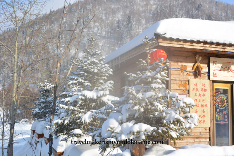 Daytime: A red lantern dangles from the roof of a timber house with snow covered trees in front and forested mountains in the back.