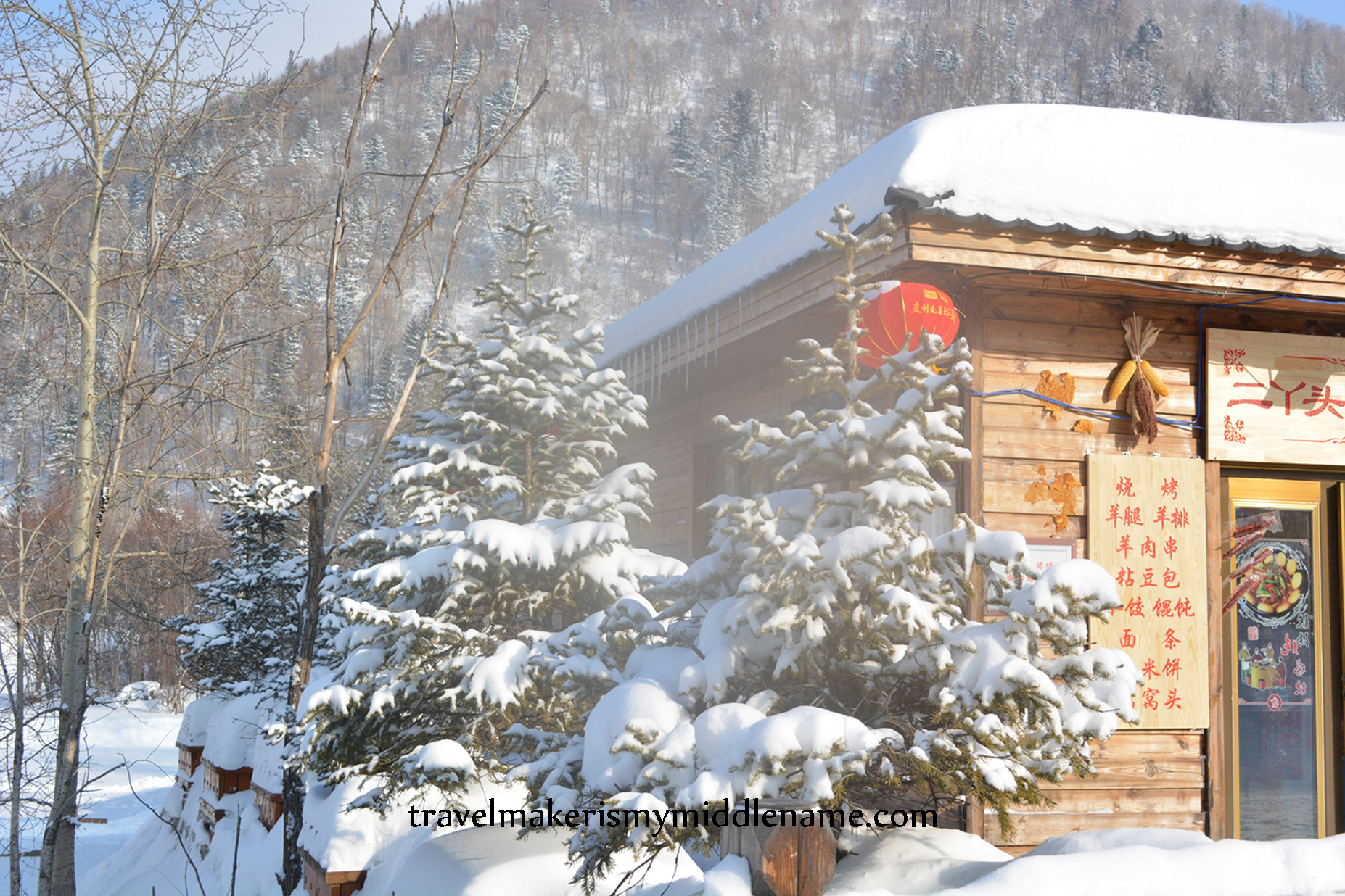 DSC_1225-WM Daytime: A red lantern dangles from the roof of a timber house with snow covered trees in front and forested mountains in the back.