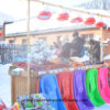 A street food vendor selling hot roasted sweet potato, corn, eggs, potatoes, traditional red sausages, sticky rice steamed buns, and varieties of frozen fruit, and colourful plastic toboggans placed neatly in front of the food stand. A woman wears a traditonal North Eastern peony floral patterned coat, the urn is covered in a layer of cloth with the same pattern.