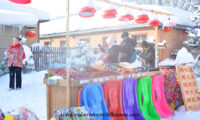 A street food vendor selling hot roasted sweet potato, corn, eggs, potatoes, traditional red sausages, sticky rice steamed buns, and varieties of frozen fruit, and colourful plastic toboggans placed neatly in front of the food stand. A woman wears a traditonal North Eastern peony floral patterned coat, the urn is covered in a layer of cloth with the same pattern.