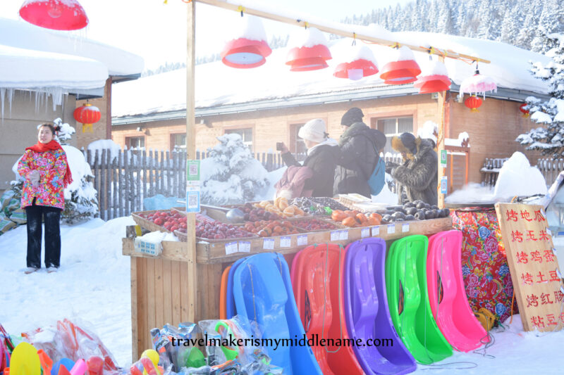 A street food vendor selling hot roasted sweet potato, corn, eggs, potatoes, traditional red sausages, sticky rice steamed buns, and varieties of frozen fruit, and colourful plastic toboggans placed neatly in front of the food stand. A woman wears a traditonal North Eastern peony floral patterned coat, the urn is covered in a layer of cloth with the same pattern.