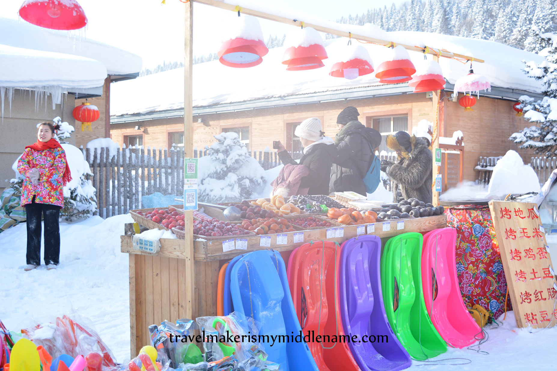 DSC_1243-WM A street food vendor selling hot roasted sweet potato, corn, eggs, potatoes, traditional red sausages, sticky rice steamed buns, and varieties of frozen fruit, and colourful plastic toboggans placed neatly in front of the food stand. A woman wears a traditonal North Eastern peony floral patterned coat, the urn is covered in a layer of cloth with the same pattern.