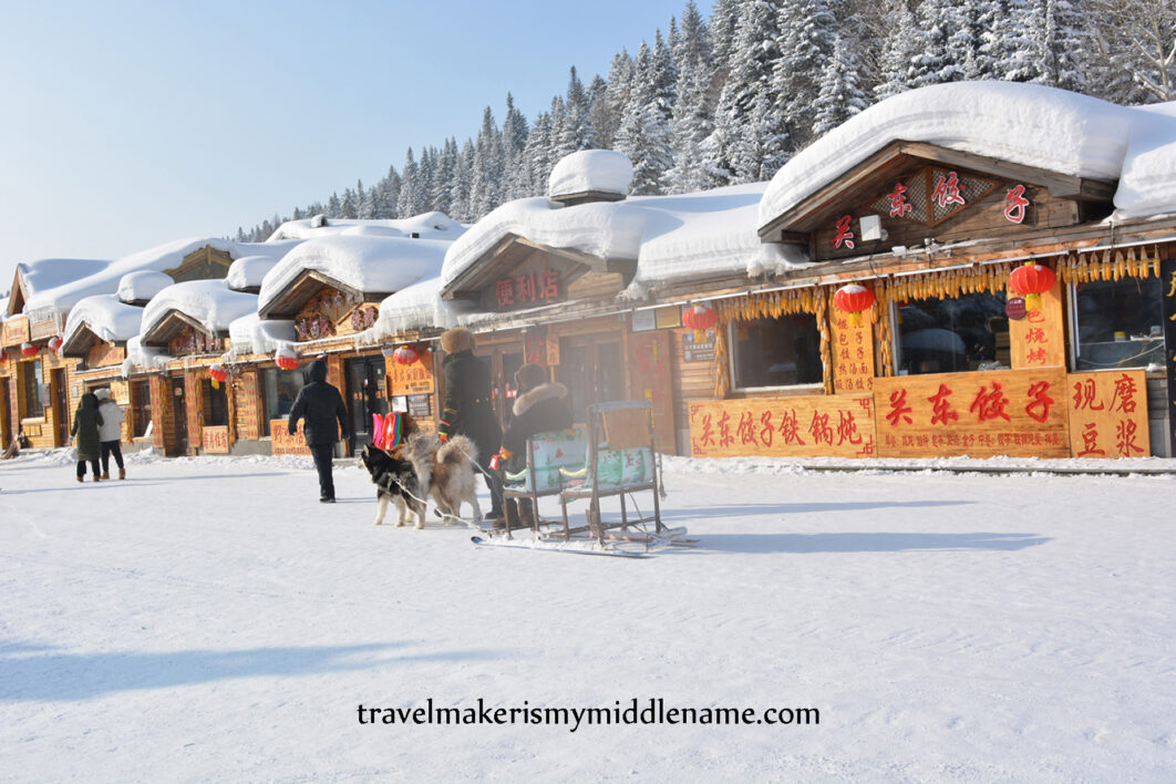 Daytime: A person rides a dog sled guided by another person through the main street in Snow Town, lined with timber style restaurants.