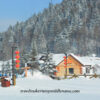 Daytime: A timber building covered in snow on a white, snow covered ground, with several red lanterns hanging in a column from street lamps and a forested mountain dusted with snow in the background.