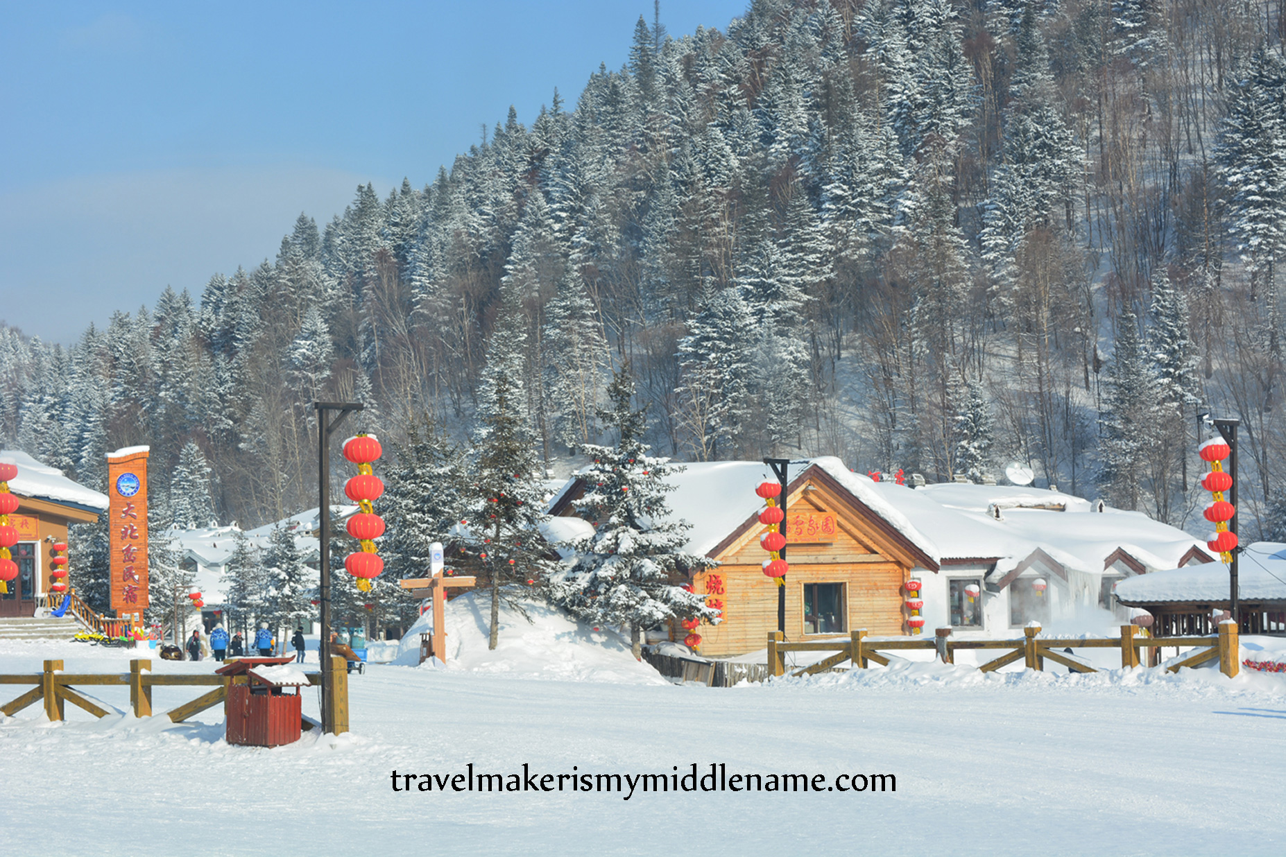 DSC_1250-WM Daytime: A timber building covered in snow on a white, snow covered ground, with several red lanterns hanging in a column from street lamps and a forested mountain dusted with snow in the background.