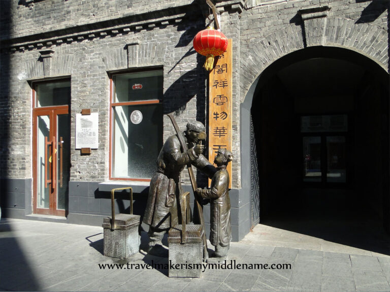 Statues of a child and a pedder selling toys from boxes carried on a stick in an alleway in the old town of Harbin. A red lantern hangs outside a wall of grey stone.