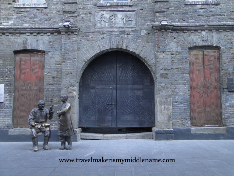 Statues of a person watching man playing the Er Hu in an alleway in the old town of Harbin. A red lantern hangs outside a wall of grey stone.