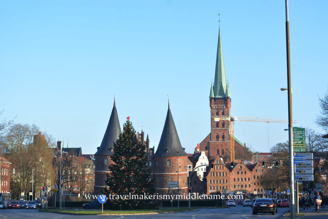 Lübeck street scenery showing the Holstentor and the Marien church in the background, and a large decorated Christmas tree in the front. Traffic is seen on the right. 