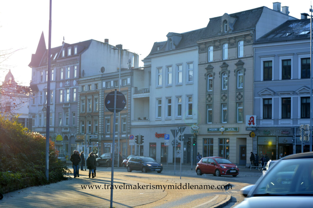 Day time: On the left: Two people walking along a wide, curved neatly paved path next to a road on a sunny winter's day on the streets of Lübeck. In the background are European style buildings. Cars drive around the curve on the road.