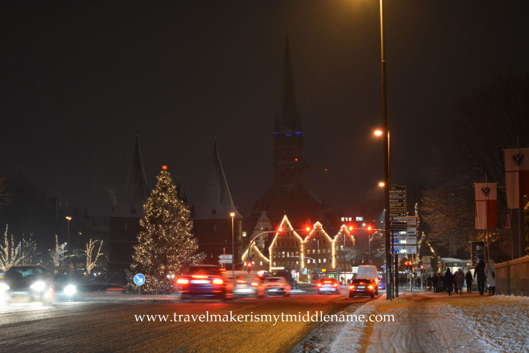 Lübeck in the middle of December at night with snow covered footpaths on the right, and traffic on the road in the middle. A lit up Christmas tree is in the background to the left, and a building outline is lit up with lights.