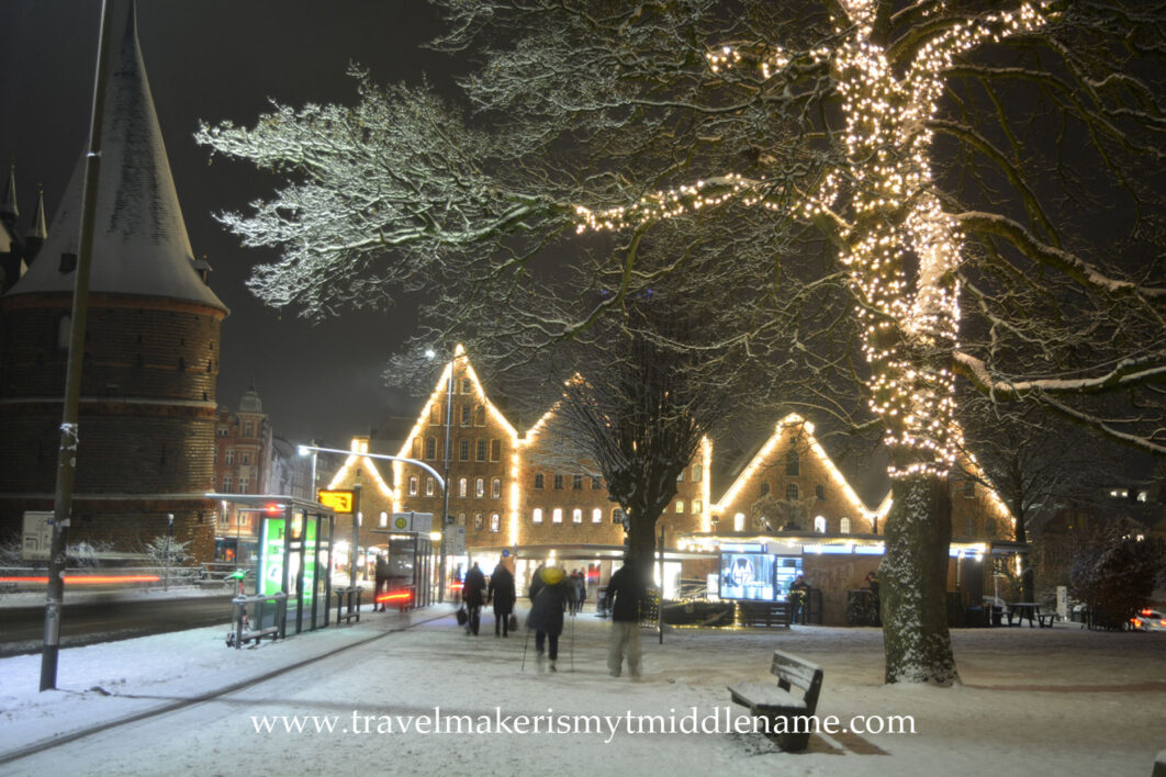 Snow covered steets of Lübeck next to the Holsten Gate in the middle of December at night. A large tree on the right has the tree trunks covered in lights.