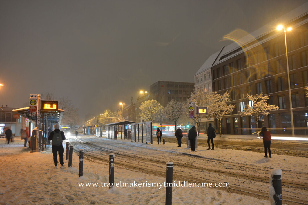 Snow covered grounds, roads and buldings at a bus stop in Lübeck in the middle of December at night. 