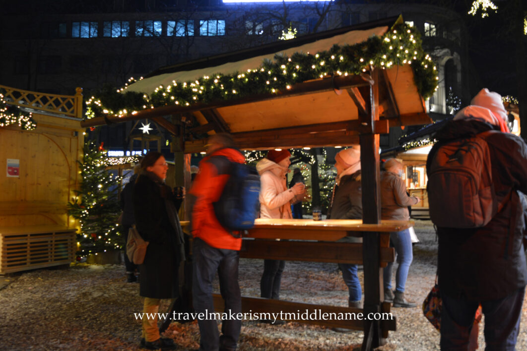 People standing at outdoor dining tables covered with a roof and decorated with lights and tinsel to eat their food at a Christmas market in Kiel, Germany