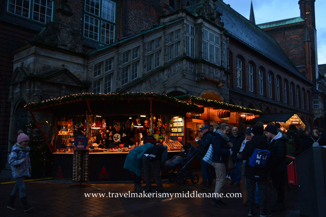 A stall selling wooden toys at the Chrismas market in Lübeck at dusk. The orange glow of the stall looks welcoming. Two parents and a child in a stroller is looking at the stall. A child on the left walks towards the right. A group of people or on the right. The stall is set in front of a medieval style church with impressive stone structures and windows.