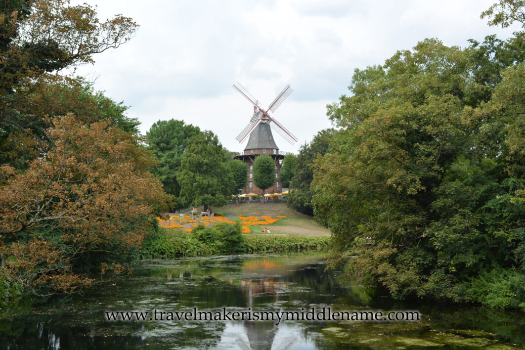 A view of the windmill atop a small grassy hill on the other side of a body of water with trees on either side during the day at the Wallanlagen park in Bremen, Germany.  