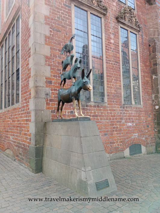 The Musicians of Bremen statue in Bremen, Germany during the day. From top to bottom in increasing size: A rooster stands on a cat, which stands on a dog, which stands on a donkey, all of which is on a skewed concrete trapezoidal prism.
